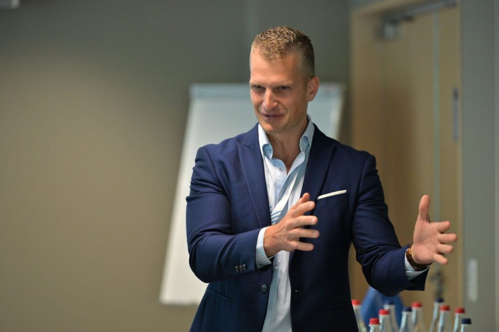A man in a blue suit gestures with his hands while speaking in an office setting, with a flipchart and bottles on a table in the background.