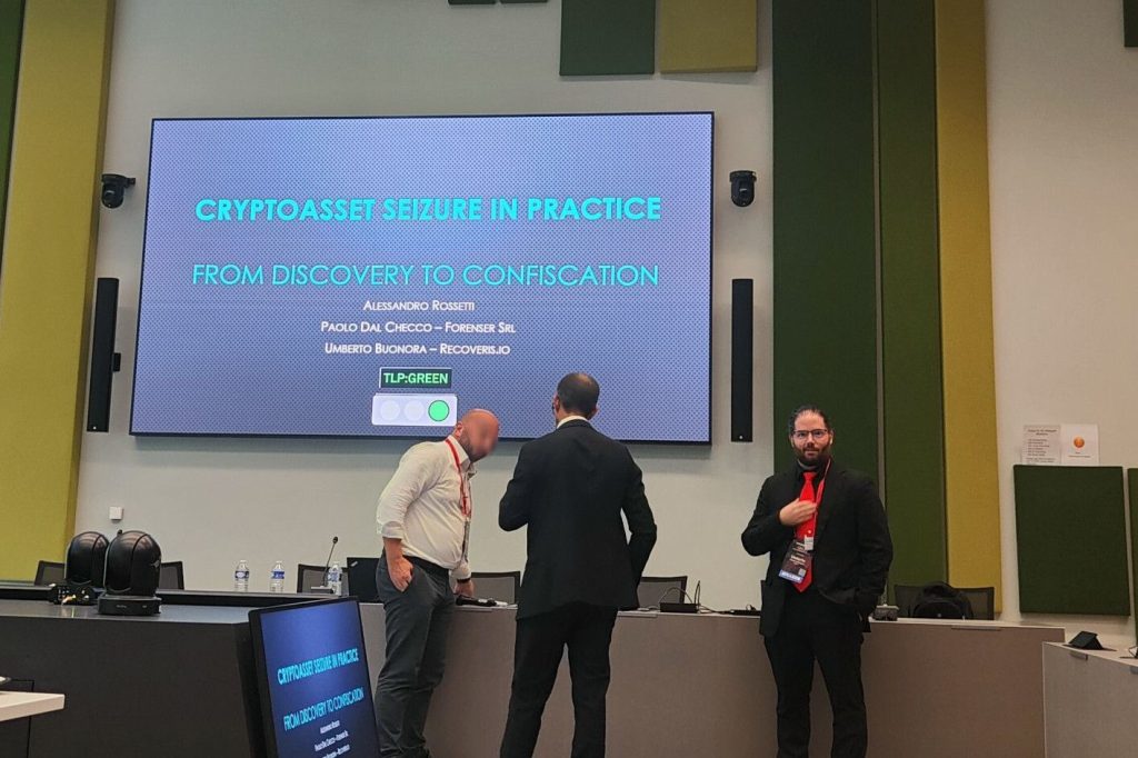 Three men in business attire stand and talk in front of a large screen displaying a presentation titled CRYPTOASSET SEIZURE IN PRACTICE: FROM DISCOVERY TO CONFISCATION in a modern conference room.