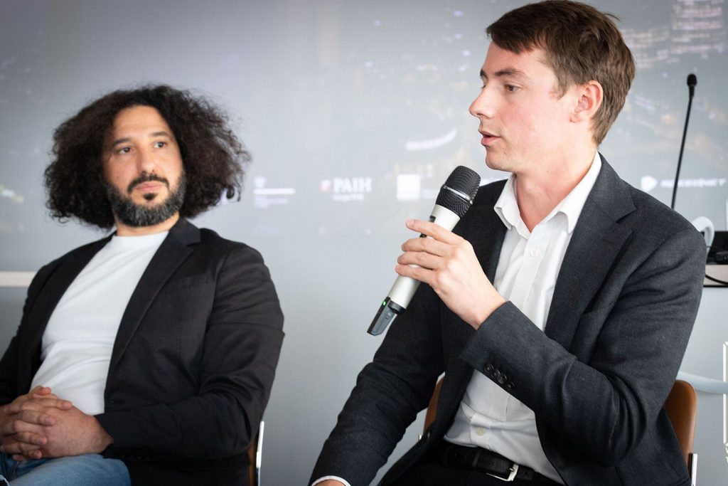 Two men in suits sit on a panel. One man with curly hair and a beard listens attentively, while the other, holding a microphone, speaks. Both wear dark blazers and light shirts. A blurred screen is in the background.