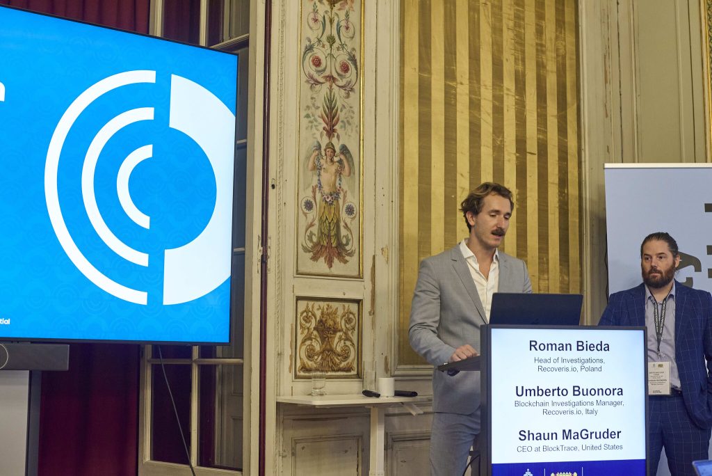 Two men in suits stand at a podium, one speaking into a microphone. A screen displays circular graphics on the left, and a monitor lists names and titles: Roman Bieda, Umberto Buonora, and Shaun MaGruder. Ornate interior walls are visible.