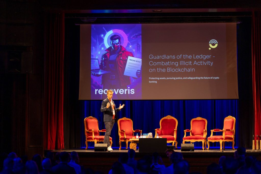 A man in a suit speaks onstage with red chairs and a table beside him. Behind him, a screen displays a graphic and text about Guardians of the Ledger – Combating Illicit Activity on the Blockchain.