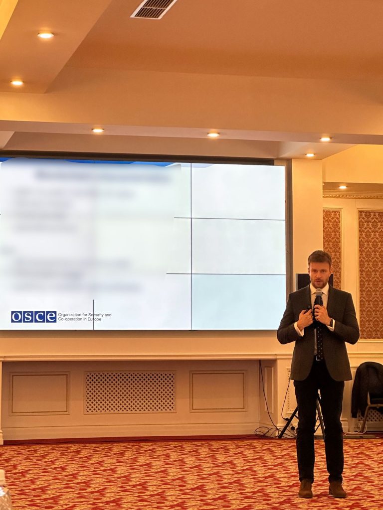 A man in a suit stands speaking into a microphone in a conference room, with a large presentation screen behind him displaying the OSCE logo and blurred text. The room has patterned carpet and decorative walls.
