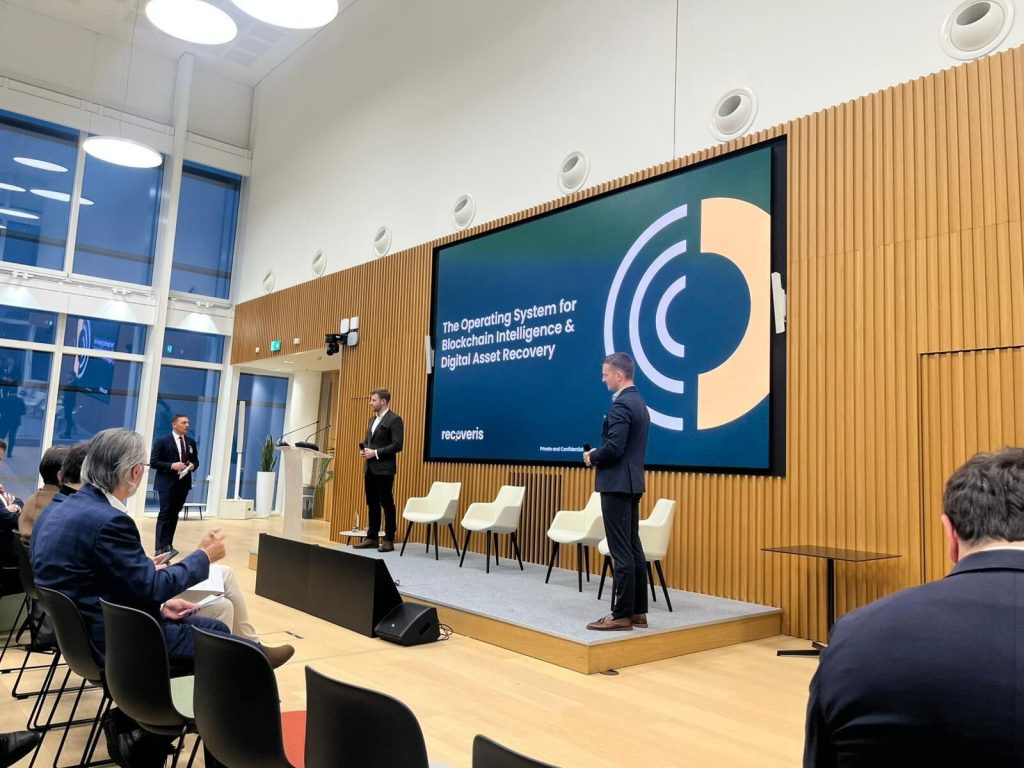 A speaker stands on stage in front of a large screen displaying The Operating System for Blockchain Intelligence & Digital Asset Recovery as an audience watches in a modern conference room with wooden paneling and large windows.