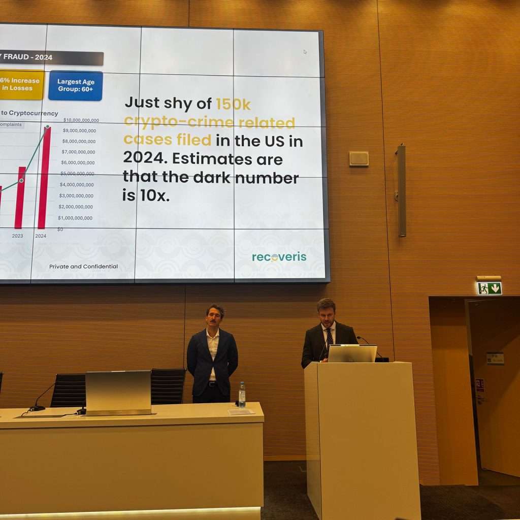 Two men in business attire stand at the front of a conference room. A presentation slide behind them highlights crypto-crime statistics, stating 15k crypto-crime related cases filed in the US in 2024 with related charts and the recoveris logo.