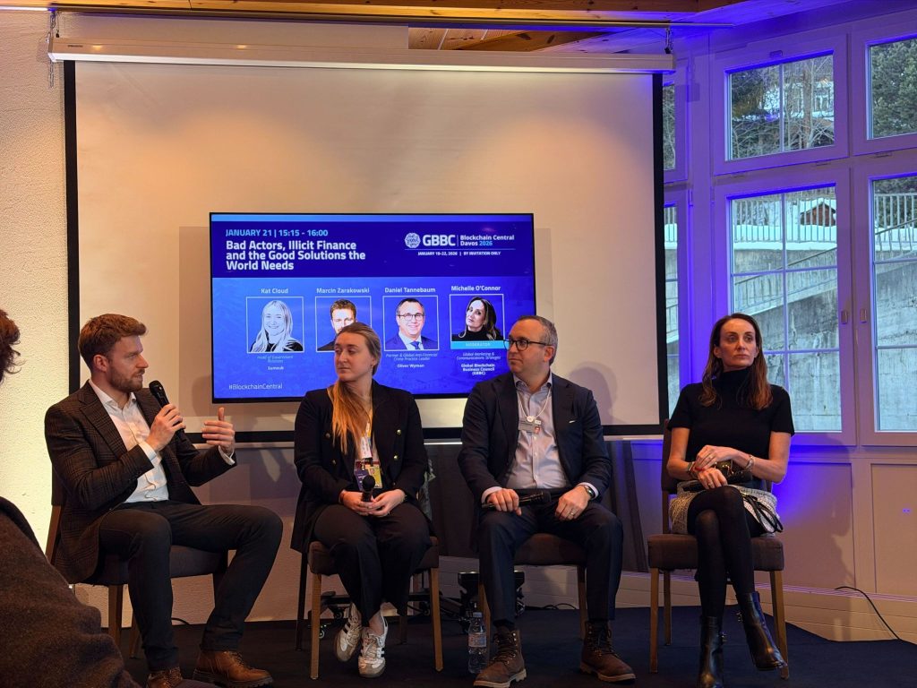 Four panelists sit on chairs in front of a screen displaying event details about Bad Actors, Illicit Finance and the Good Solutions the World Needs. One man speaks while the others listen in a bright room with large windows.