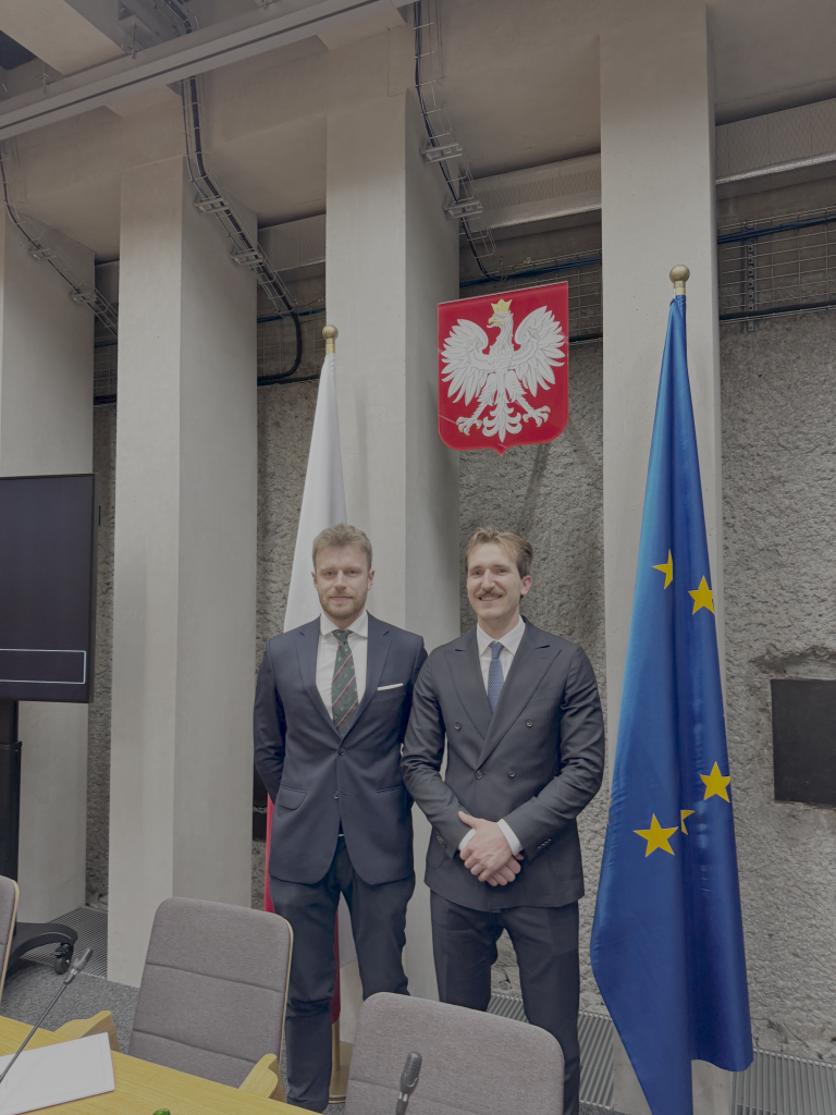 Two men in suits stand side by side indoors, in front of the Polish coat of arms, with the flags of Poland and the European Union visible behind them.