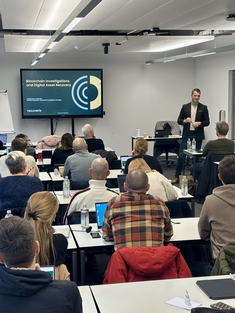 A man stands and speaks in front of a classroom of seated adults. Behind him, a presentation slide reads “Blockchain Investigations and Digital Asset Recovery.” Attendees face forward, some using laptops and tablets.