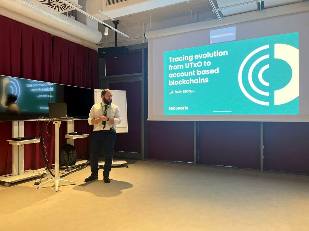 A man stands in a conference room giving a presentation. Behind him is a large screen displaying a slide titled Tracing evolution from UTxO to account based blockchains, with the company name recoveris at the bottom.